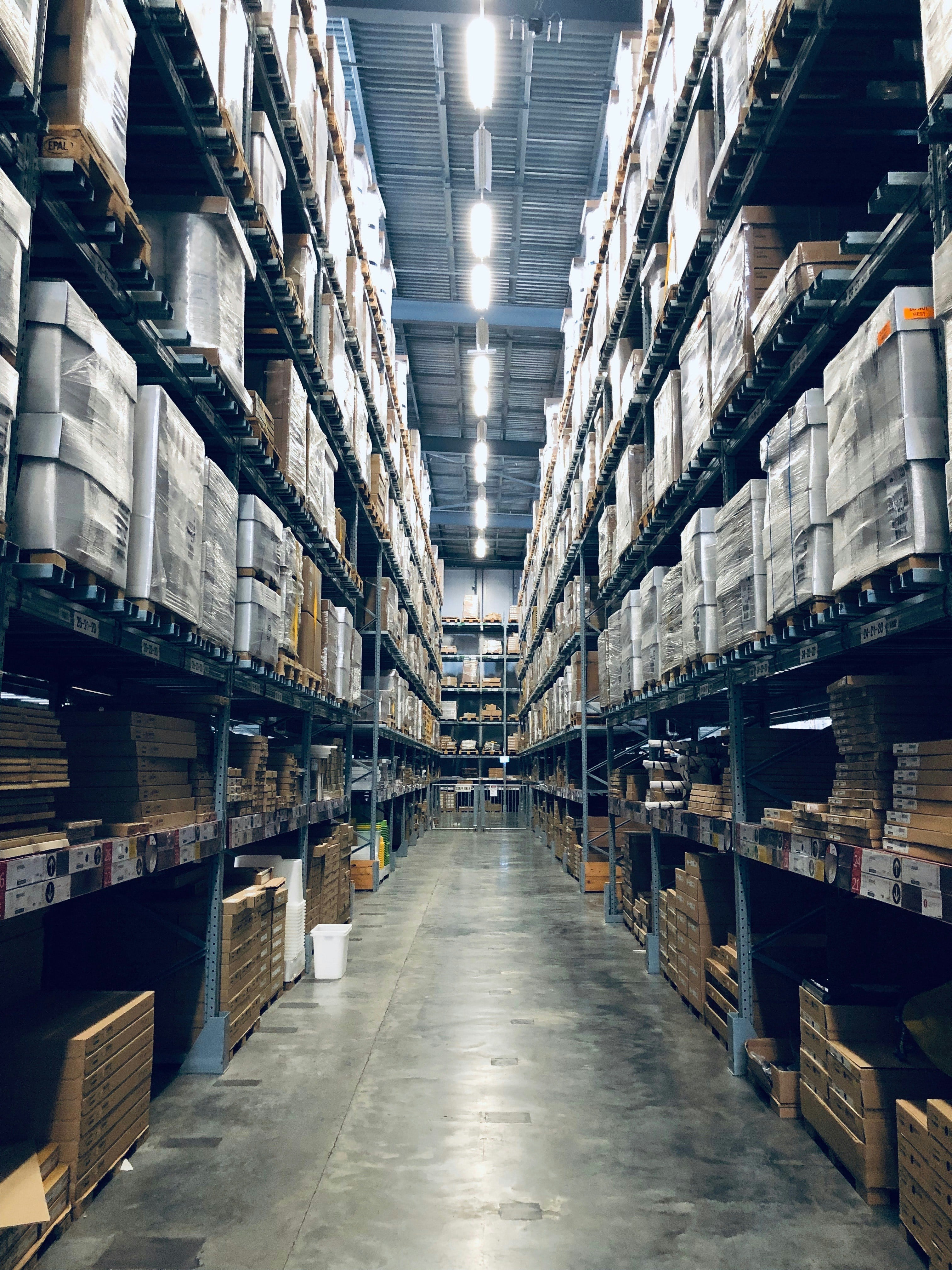 Long aisle of a distribution warehouse, tall pallet racking stacked with boxes receding toward the far wall