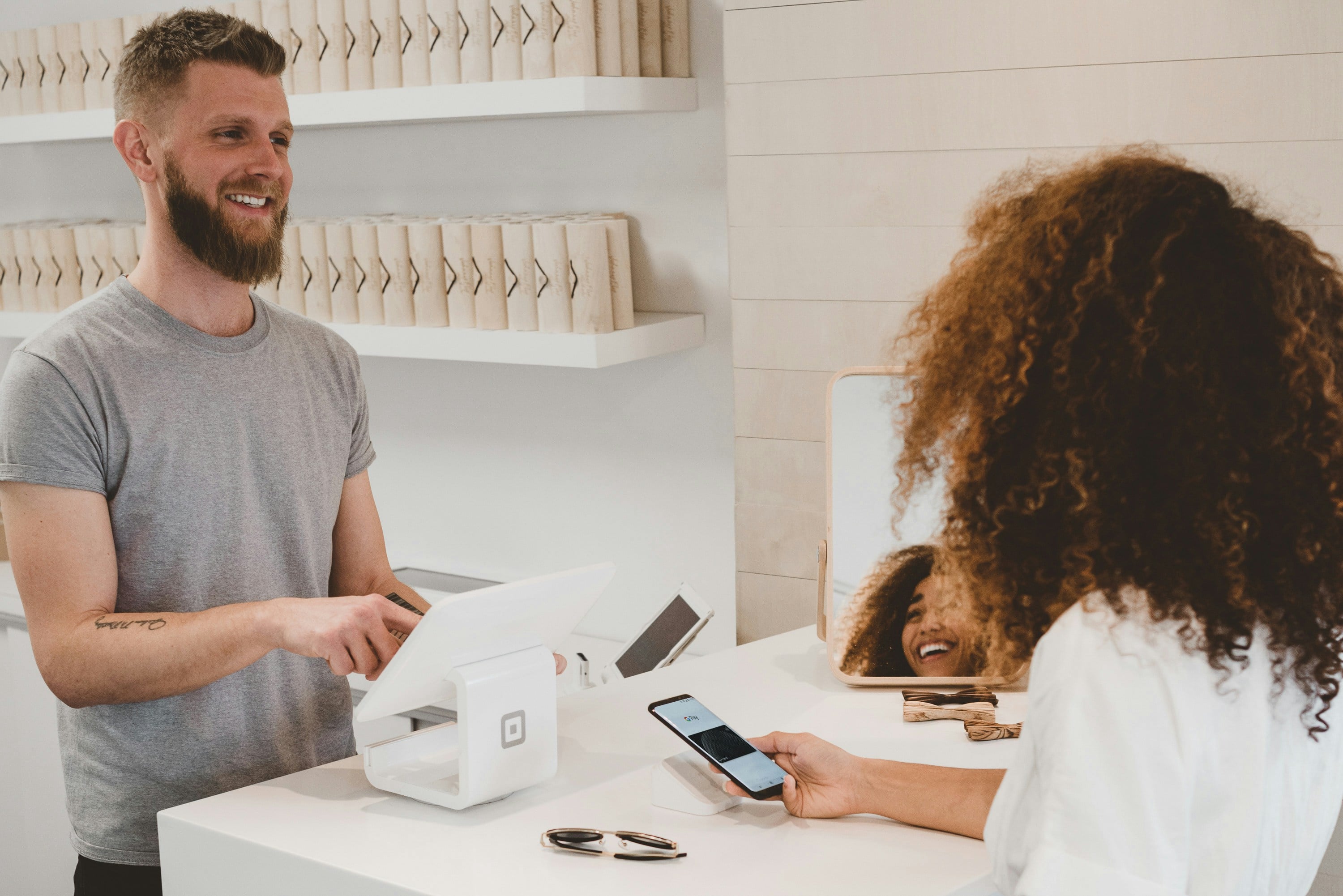 Two people talking across a shop counter in daylight, one leaning in while the other checks a tablet